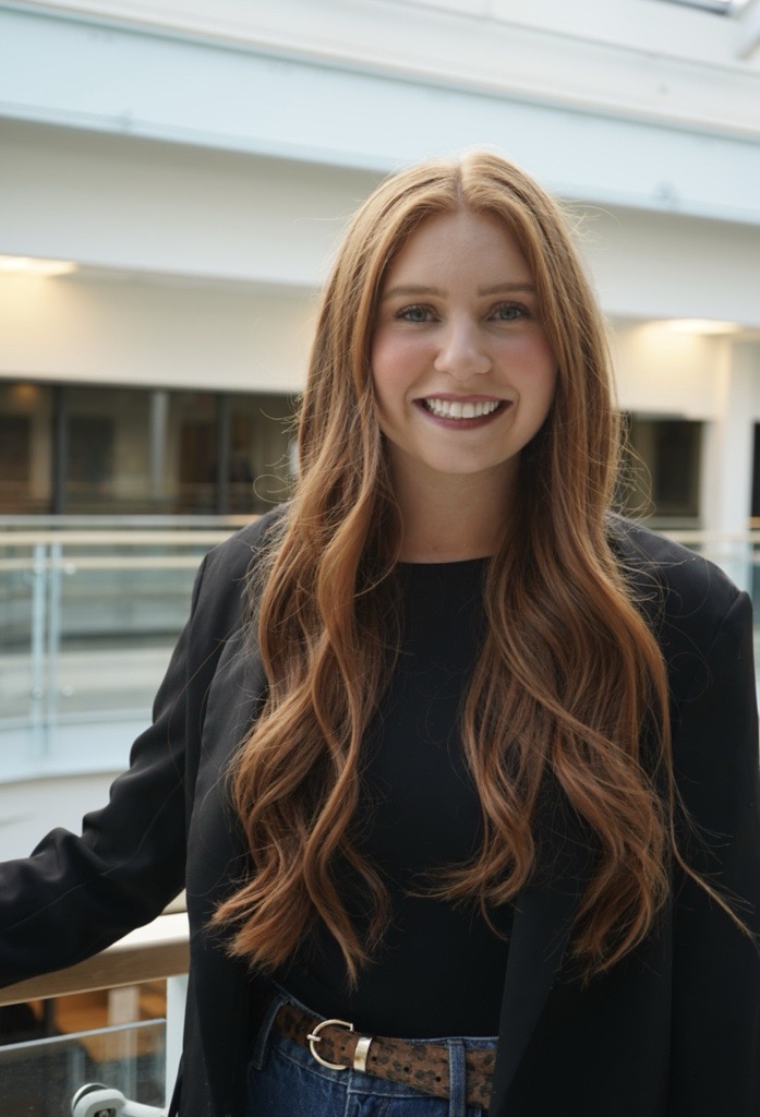 Olivia is a white woman with long red hair. She wears a black shirt and blazer.